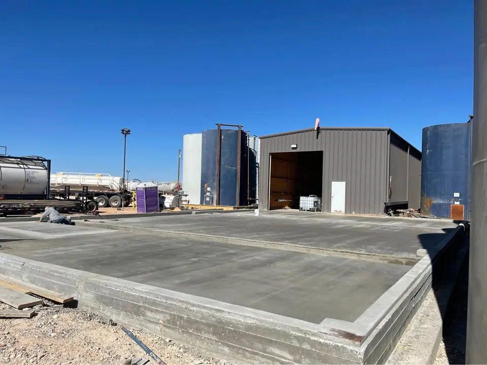 In Buckeye, AZ, a construction site showcases a freshly poured concrete foundation by Bucks Concrete Craft. Behind it stands a gray industrial building with an open entrance, surrounded by large storage tanks and a parked tanker truck under the clear blue sky.