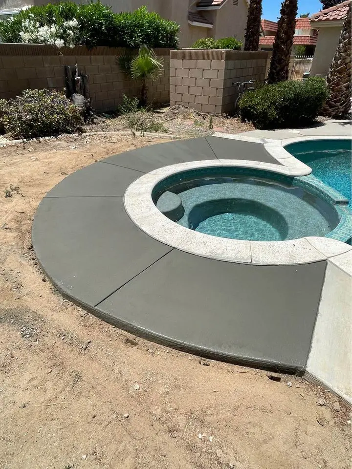 A serene backyard scene in Buckeye, Arizona features an in-ground pool with a circular hot tub partially surrounded by fresh concrete expertly laid by a skilled pool deck contractor. Nearby, a small garden and palm trees add to the tranquil atmosphere.