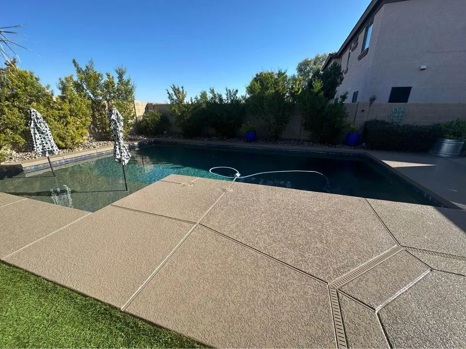 A backyard pool with a concrete deck crafted by expert pool deck contractors features two closed umbrellas. The inviting oasis, surrounded by lush greenery and a tan wall, basks under a clear blue sky.