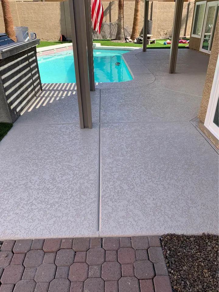 A backyard scene in Buckeye, Arizona, showcases a swimming pool with a concrete patio crafted by a skilled pool deck contractor. A small flag flutters gently amid patio furniture and trees in the background. The foreground features a harmonious blend of concrete and brick flooring.