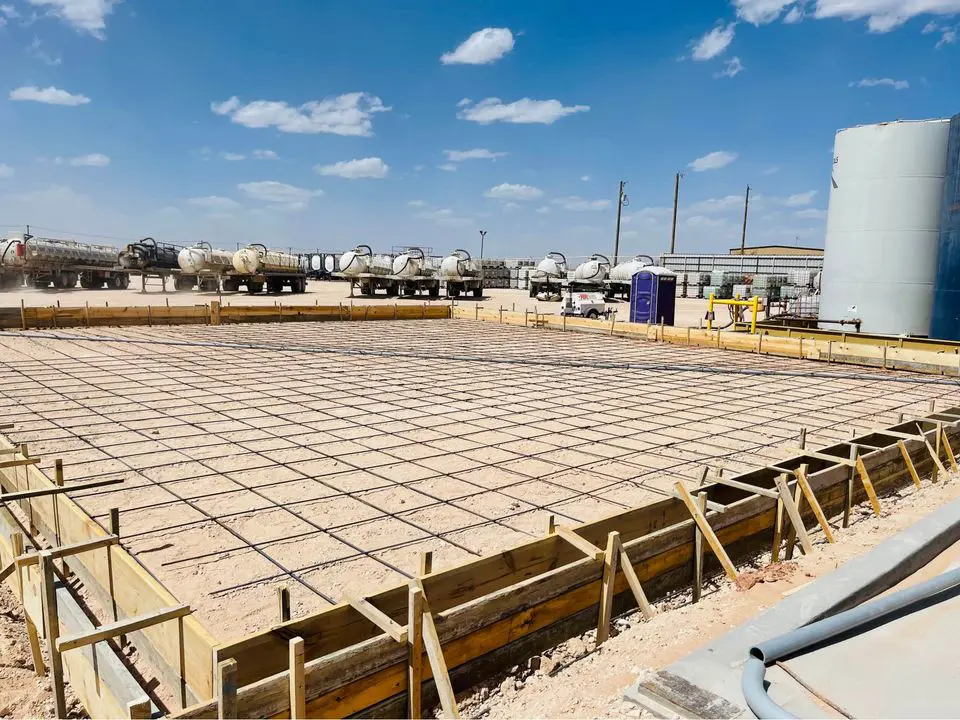 In Buckeye AZ, a construction site features a large rectangular foundation in progress, surrounded by wooden formwork and steel rebar. Concrete contractors are on hand as several trucks are parked in the background under a clear blue sky with clouds.