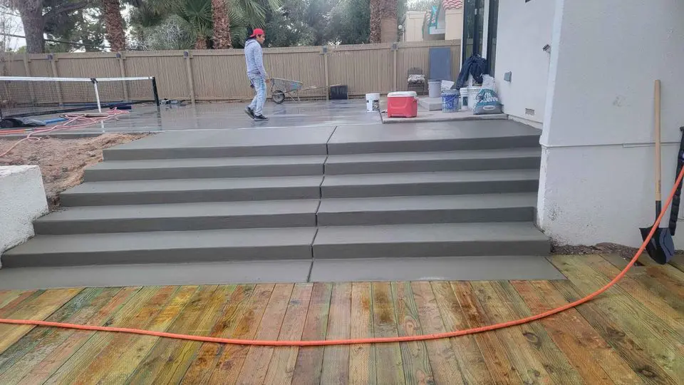 A person in a gray outfit and red cap walks on a wet concrete surface near large stairs, likely crafted by skilled concrete contractors. Construction tools and materials are visible. The area includes a wooden deck, a tennis net, and a few trees in the background.