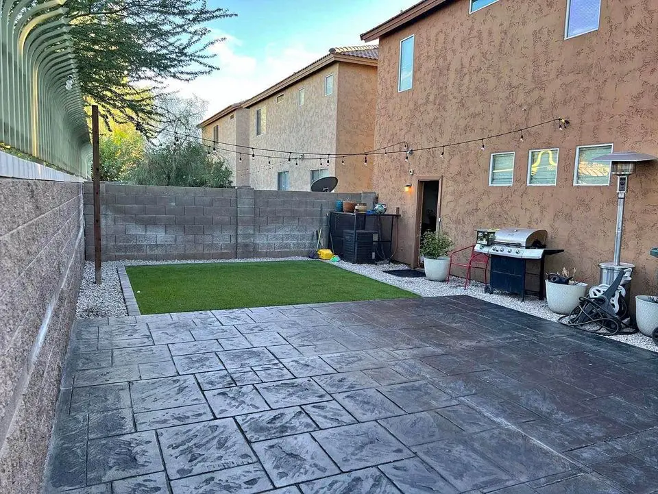 The backyard features a spacious stone patio, ideal for surprise gatherings, with a small grass area and string lights overhead. Enclosed by a stone wall, it includes a barbecue grill and potted plants to the right. Two-story houses envelop this cozy retreat.