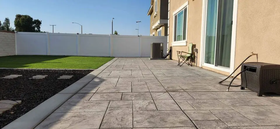 In the backyard, a spacious stone-tiled patio merges seamlessly with a lush green lawn and a white fence. A small folding chair and box sit near the house. The sun casts shadows on the tiles, while landscaping stones enhance the charm on the left—perfect for Goodyear AZ residents seeking concrete services.