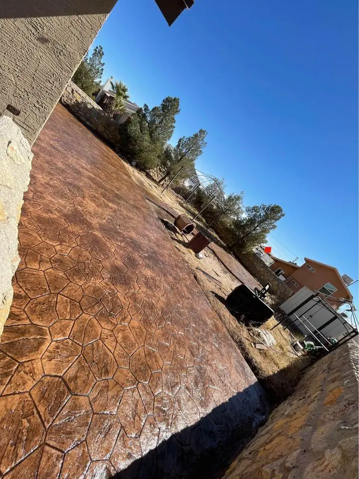 A backyard in Buckeye AZ showcases a concrete patio with a hexagonal pattern crafted by skilled concrete contractors. Nestled beside a stone wall, it offers views of trees and a wheelbarrow under the clear blue sky.