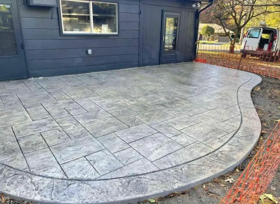 A freshly poured concrete patio with a stamped tile pattern graces a Buckeye backyard. The patio curves elegantly, bordered by orange safety fencing—a hallmark of expert Concrete Patio Contractors at work. A van is parked nearby a dark building, hinting at the surprise transformation in progress.