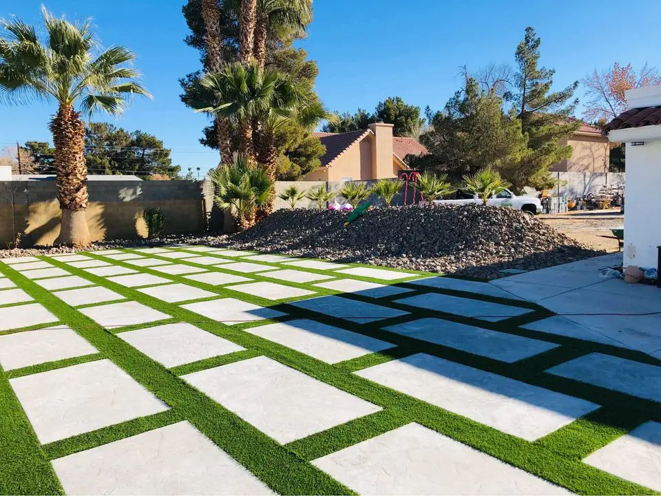 A backyard in Buckeye, AZ features a grid of rectangular concrete slabs interspersed with lush artificial grass. Palm trees and a rock pile adorn the backdrop under a clear blue sky, while on the right, part of the house and expertly crafted concrete driveway are visible.