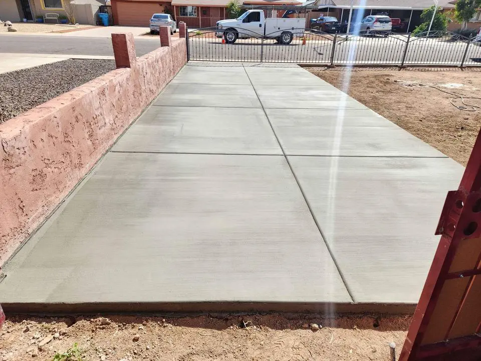 A freshly poured concrete driveway, skillfully crafted by local concrete contractors in Goodyear, AZ, boasts a smooth finish. It's bordered by a reddish wall on the left and a metal fence and gate at the end, with a pickup truck visible on the street in the background.