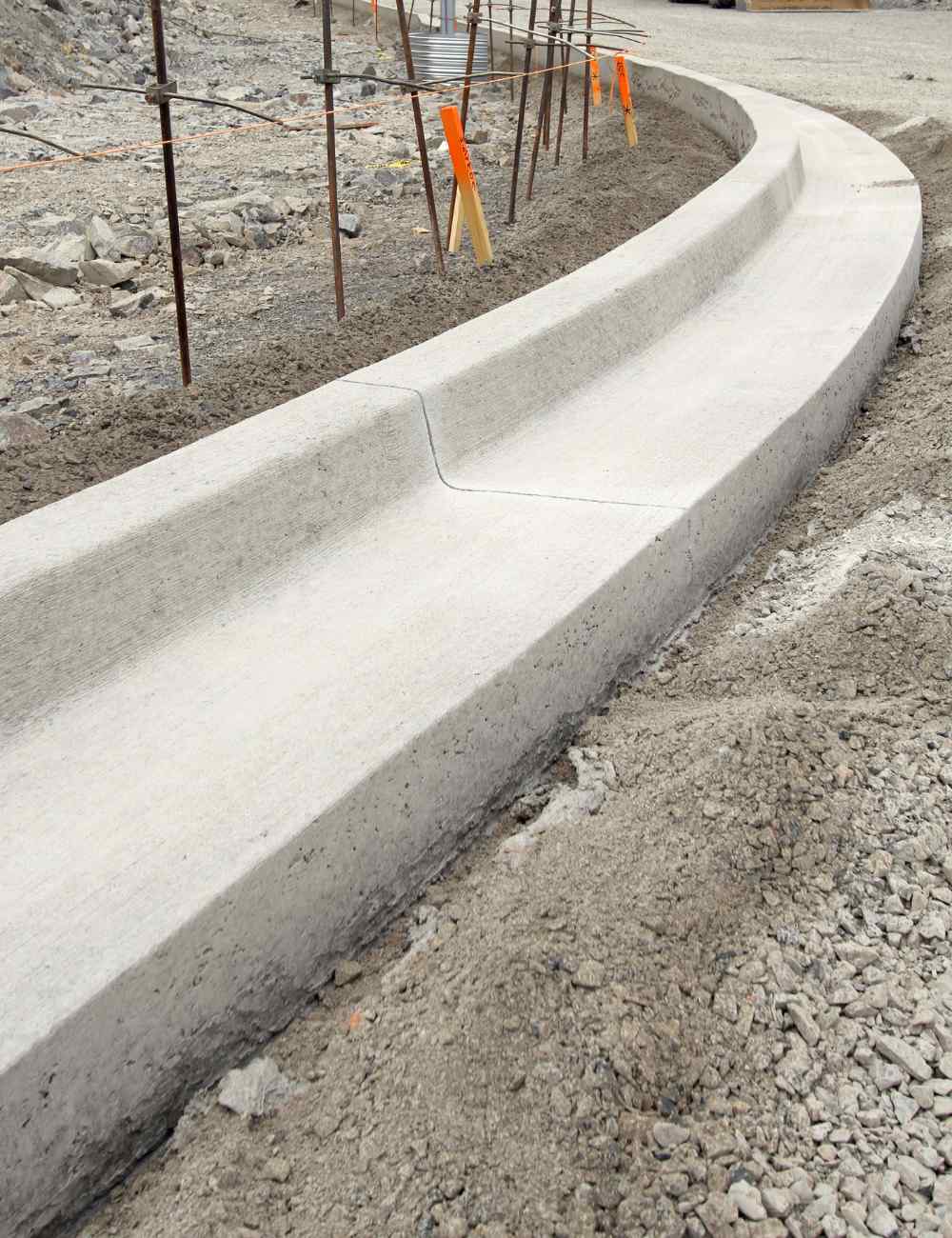 A newly constructed concrete curb on a road under construction curves to the right, surrounded by loose gravel and construction materials. Orange markers line the path, while scaffolding looms in the background. Skilled concrete contractors are also working on nearby driveways and patios.